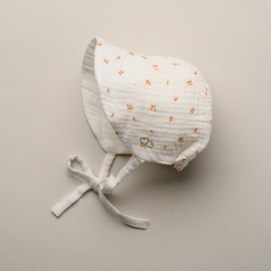May include: A white baby bonnet with a cherry pattern. The bonnet is made of a soft, textured fabric and features a small bow at the chin. The cherries are pink and green. The bonnet has a small embroidered design.
