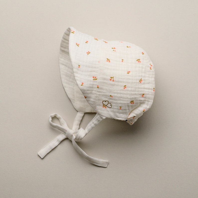 May include: A white baby bonnet with a cherry pattern. The bonnet is made of a soft, textured fabric and features a small bow at the chin. The cherries are pink and green. The bonnet has a small embroidered design.