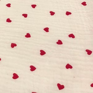 May include: A white fabric with a pattern of scattered red hearts. The hearts are evenly spaced across the surface, creating a playful and romantic design. The fabric has a slightly textured appearance.