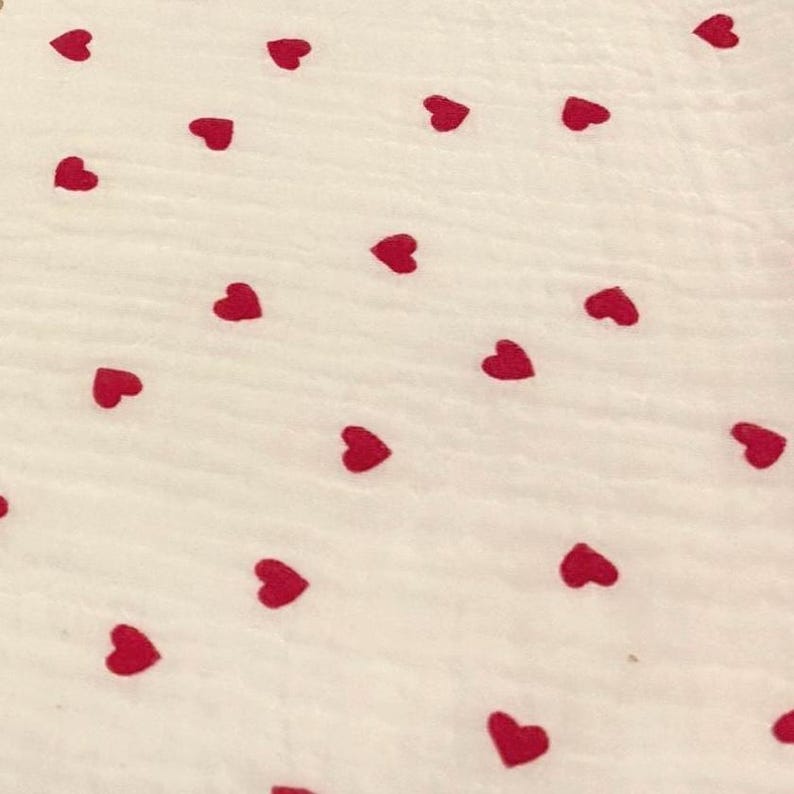 May include: A white fabric with a pattern of scattered red hearts. The hearts are evenly spaced across the surface, creating a playful and romantic design. The fabric has a slightly textured appearance.