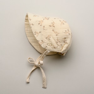 May include: A cream-colored baby bonnet with a floral pattern of brown branches and pink berries. The bonnet has a soft, textured appearance and is tied with cream-colored fabric ties. The bonnet is designed to protect the baby's head from the sun.