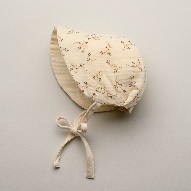 May include: A cream-colored baby bonnet with a floral pattern of brown branches and pink berries. The bonnet has a soft, textured appearance and is tied with cream-colored fabric ties. The bonnet is designed to protect the baby's head from the sun.