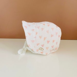 May include: A white baby bonnet with a pattern of light pink hearts. The bonnet has a rounded shape with a tie closure and a small gold cloud logo. The fabric appears soft and lightweight, suitable for a baby.