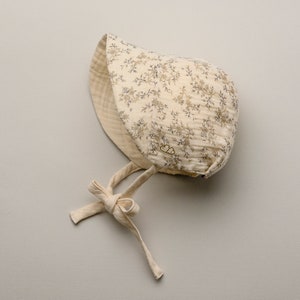 May include: A cream-colored baby bonnet with a floral pattern in shades of brown and blue. The bonnet has a soft, quilted texture and ties under the chin with cream-colored fabric ties. A small logo is visible on the side.