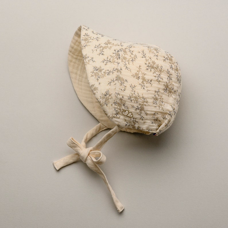 May include: A cream-colored baby bonnet with a floral pattern in shades of brown and blue. The bonnet has a soft, quilted texture and ties under the chin with cream-colored fabric ties. A small logo is visible on the side.