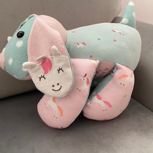 May include: A soft, plush toy shaped like a dog with a unicorn face. The toy is made of blue and pink fabric with a white unicorn pattern. The toy has a white unicorn face with pink cheeks and eyelashes. The toy is sitting on a light grey sofa.