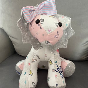 May include: A gray and pink stuffed lion with a blue and pink bow. The lion has a white body with a pink heart and a unicorn design. The lion's paws have a gray and white fabric with a heart and shoe design. The lion's paws have the text "I love you Dad" on them.