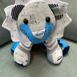 May include: A handmade stuffed lion toy made from repurposed fabric. The lion is blue, gray, and white with a white mane. The fabric has a pattern of cars and dogs. The lion has black button eyes and a tag that says "Handsome Guy".