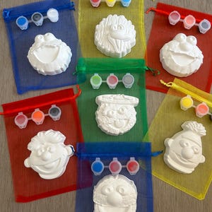 May include: Seven colourful mesh bags containing white ceramic character figurines and paint pots. The characters include Cookie Monster, Elmo, Big Bird, Abby Cadabby, Oscar the Grouch, and Bert and Ernie.