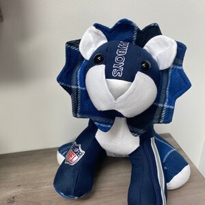 May include: Blue and white plush lion with a plaid mane and the word "COWBOYS" embroidered on the chest. The lion is wearing a blue and white NFL patch on its leg.