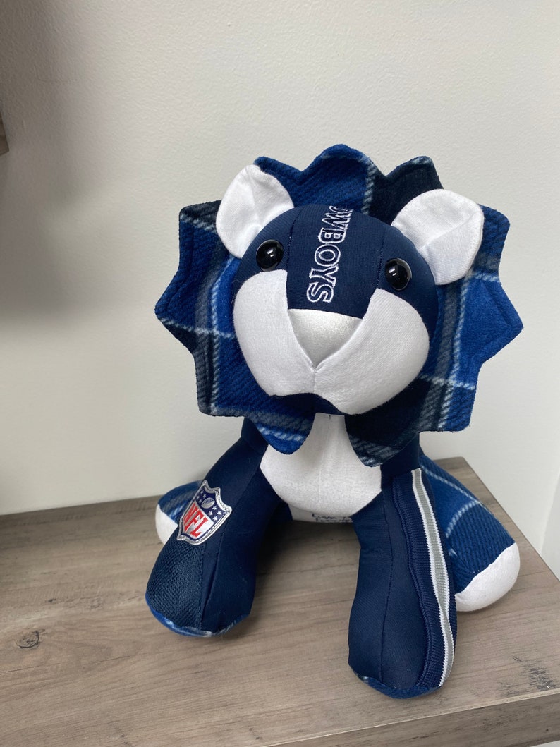May include: Blue and white plush lion with a plaid mane and the word "COWBOYS" embroidered on the chest. The lion is wearing a blue and white NFL patch on its leg.