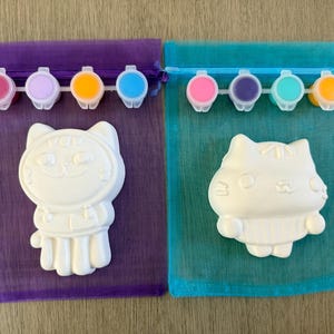 Puede incluir: Dos kits de pintura de gatos. Cada kit incluye una figura de gato de cer&aacute;mica blanca, una peque&ntilde;a paleta de pintura con varios colores y una bolsa transparente con cord&oacute;n. Los kits est&aacute;n sobre una superficie de madera marr&oacute;n claro.