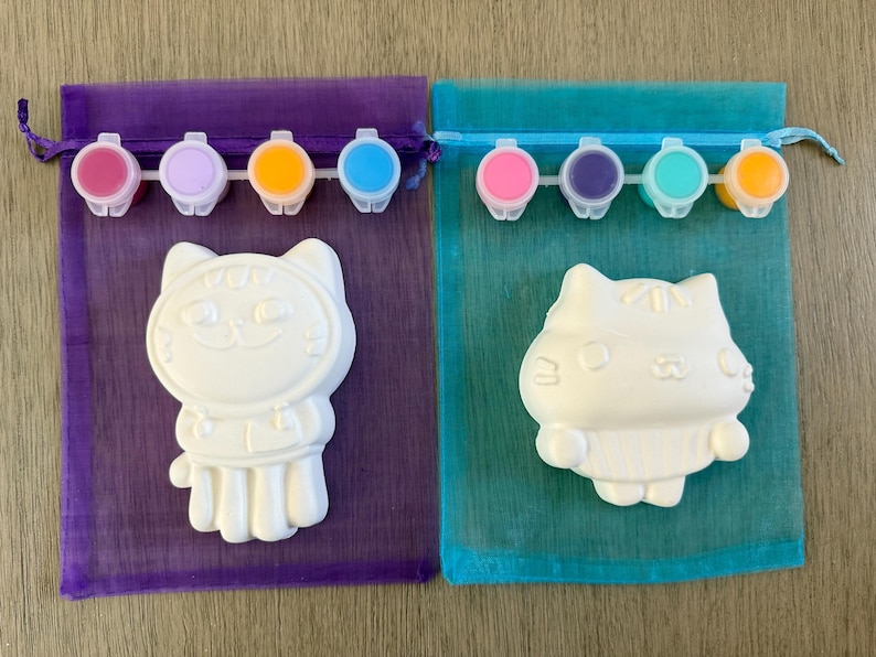 Puede incluir: Dos kits de pintura de gatos. Cada kit incluye una figura de gato de cer&aacute;mica blanca, una peque&ntilde;a paleta de pintura con varios colores y una bolsa transparente con cord&oacute;n. Los kits est&aacute;n sobre una superficie de madera marr&oacute;n claro.