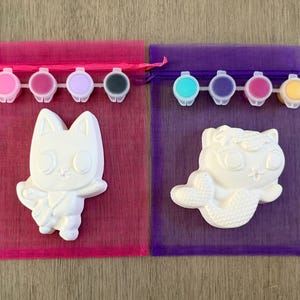 Puede incluir: Dos kits de pintura para hacer uno mismo, cada uno con una figura de cer&aacute;mica blanca y botes de pintura. Un kit presenta una figura de gato, el otro un gato sirena. Los kits est&aacute;n en bolsas de malla rosa y morada con cierres de cord&oacute;n.