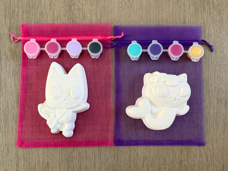Puede incluir: Dos kits de pintura para hacer uno mismo, cada uno con una figura de cer&aacute;mica blanca y botes de pintura. Un kit presenta una figura de gato, el otro un gato sirena. Los kits est&aacute;n en bolsas de malla rosa y morada con cierres de cord&oacute;n.