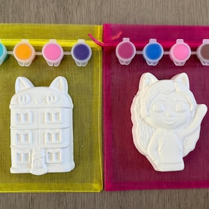 Puede incluir: Dos kits para pintar. Uno presenta un edificio blanco con forma de gato, el otro un personaje de dibujos animados blanco. Cada kit incluye un juego de botes de pintura de varios colores y una bolsa de malla.