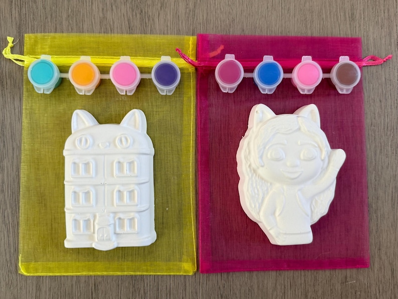 Puede incluir: Dos kits para pintar. Uno presenta un edificio blanco con forma de gato, el otro un personaje de dibujos animados blanco. Cada kit incluye un juego de botes de pintura de varios colores y una bolsa de malla.