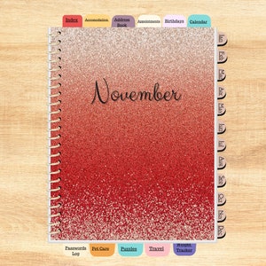 以下が含まれることがあります： 赤い金色のグリッターのノート。スパイラル製本で、1 年の各月のためのタブが付いています。表紙には「11 月」と書いてあります。