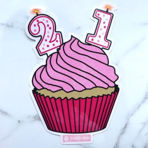 Puede incluir: Una pegatina de cupcake rosa con un remolino de glaseado rosa, un envoltorio dorado y dos velas que dicen "21". Las velas son blancas con lunares rosas. La pegatina tiene un código Spotify en la parte inferior.