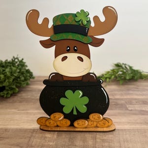 May include: A wooden St. Patrick's Day decoration featuring a moose with antlers wearing a green hat with a shamrock. The moose is peeking out of a black pot filled with gold coins and a green shamrock.