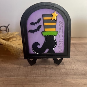 May include: A black wooden Halloween decoration featuring a witch's boot and hat with a star. The background is purple with two bat silhouettes. The text "witch please" is written vertically on the right side of the frame.
