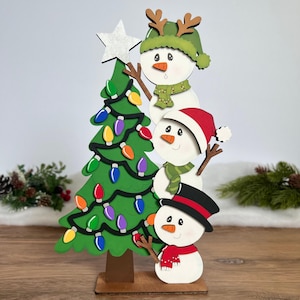 Puede incluir: Una decoración navideña de madera con un árbol verde, luces de colores y una estrella plateada en la punta. Tres muñecos de nieve apilados están al lado, con un gorro verde con astas, un gorro rojo y un sombrero de copa negro con bufandas.