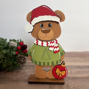 Puede incluir: Una figura de oso navide&ntilde;o de madera con gorro de Pap&aacute; Noel rojo, su&eacute;ter verde y bufanda roja y blanca. El oso sostiene un adorno rojo con un lazo dorado. La figura est&aacute; sobre una base de madera.
