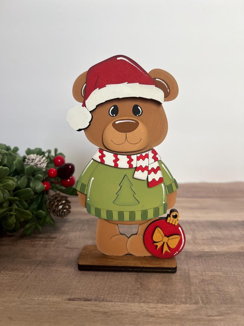 Puede incluir: Una figura de oso navide&ntilde;o de madera con gorro de Pap&aacute; Noel rojo, su&eacute;ter verde y bufanda roja y blanca. El oso sostiene un adorno rojo con un lazo dorado. La figura est&aacute; sobre una base de madera.
