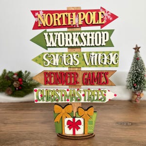 Puede incluir: Un letrero navideño de madera con cinco flechas direccionales apiladas con el texto NORTH POLE, WORKSHOP, SANTA'S VILLAGE, REINDEER GAMES y CHRISTMAS TREES. La base está decorada con tres cajas de regalo con lazos amarillos y rojos.