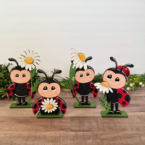 May include: Four wooden ladybug figurines with red and black bodies, black antennae, and smiling faces. Each ladybug holds a white daisy or has one on its head. They stand on green bases.