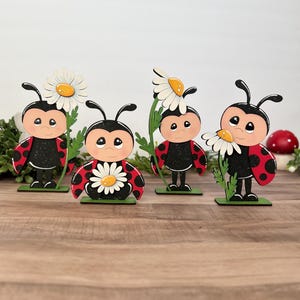 May include: Four wooden ladybug figurines with black bodies, red spotted wings, and smiling faces. Each ladybug holds or is adorned with a white daisy with a yellow center. The figurines stand on green bases, creating a whimsical garden theme.