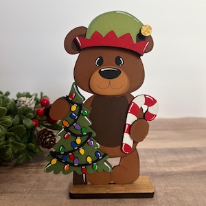 Puede incluir: Figura de oso navide&ntilde;o de madera que lleva un gorro de elfo y sostiene un &aacute;rbol de Navidad decorado y un bast&oacute;n de caramelo. El oso es marr&oacute;n con un hocico marr&oacute;n claro y est&aacute; sobre una base de madera.