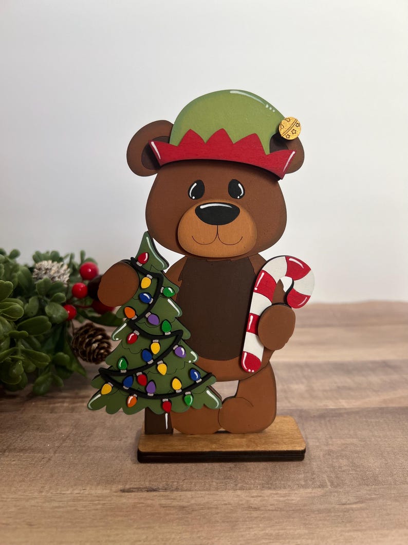 Puede incluir: Figura de oso navide&ntilde;o de madera que lleva un gorro de elfo y sostiene un &aacute;rbol de Navidad decorado y un bast&oacute;n de caramelo. El oso es marr&oacute;n con un hocico marr&oacute;n claro y est&aacute; sobre una base de madera.