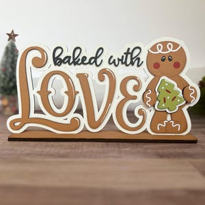 Puede incluir: Un letrero de madera con las palabras "baked with LOVE" en letras marrones y blancas. Un hombre de jengibre sosteniendo un árbol de Navidad verde está en el lado derecho del letrero. El letrero está sobre una base de madera marrón.