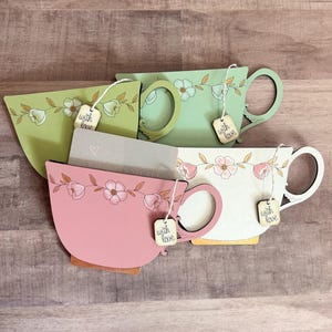 May include: Four decorative wooden tea cup cutouts in shades of green, pink, and white. Each cup features a floral design and a small wooden tag that says "with love". The cups are arranged on a wooden surface.