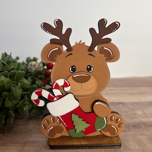 Puede incluir: Decoraci&oacute;n navide&ntilde;a de madera con un h&iacute;brido reno-oso. La figura marr&oacute;n sostiene un calcet&iacute;n rojo y verde con un dise&ntilde;o de &aacute;rbol de Navidad y un bast&oacute;n de caramelo. La figura tiene cuernos marrones y est&aacute; sobre una base de madera.