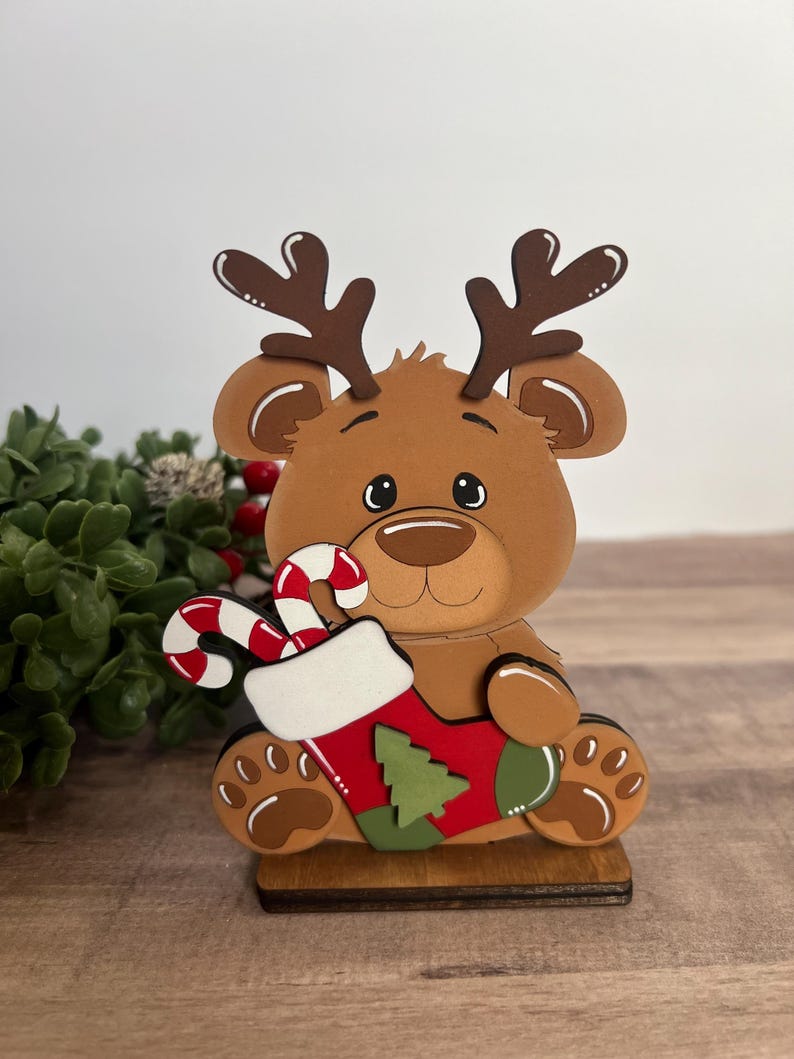 Puede incluir: Decoraci&oacute;n navide&ntilde;a de madera con un h&iacute;brido reno-oso. La figura marr&oacute;n sostiene un calcet&iacute;n rojo y verde con un dise&ntilde;o de &aacute;rbol de Navidad y un bast&oacute;n de caramelo. La figura tiene cuernos marrones y est&aacute; sobre una base de madera.