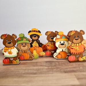 Puede incluir: Una colección de figuras de osos de madera, cada una adornada con ropa y accesorios con temática otoñal. Los osos visten suéteres, sombreros y bufandas, y sostienen calabazas. La paleta de colores incluye tonos de marrón, naranja y amarillo.
