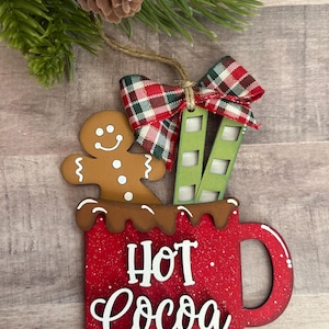 Puede incluir: Adorno navideño de madera con forma de taza roja con la inscripción "Hot Cocoa" en blanco. Un hombre de jengibre y una escalera verde y blanca están dentro de la taza. Un lazo a cuadros y cuerda completan el diseño.