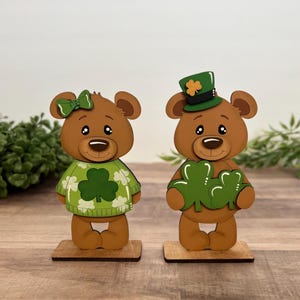 May include: Two wooden bear figurines, one wearing a green sweater with a shamrock and a green bow, the other holding shamrocks and wearing a green top hat with a shamrock. Both are on wooden bases.