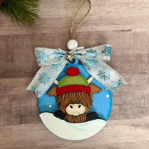 Puede incluir: Adorno navideño redondo azul con una vaca de las Tierras Altas con un gorro verde y rojo, en una escena nevada. El adorno tiene un lazo blanco con detalles de copos de nieve azules y una cuerda de yute para colgar.