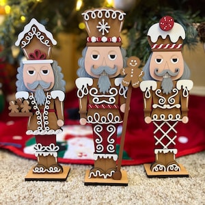 May include: Three gingerbread-themed nutcracker figurines. Each figure has a brown body with white icing details, gray hair, and a mustache. One holds cookies, another a gingerbread man, and the third has a cherry on its hat. They stand on brown bases.