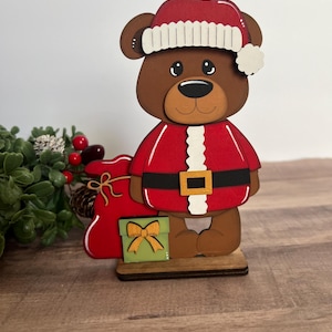 Puede incluir: Figura navide&ntilde;a de oso de madera con traje y gorro de Pap&aacute; Noel rojos con ribete blanco. El oso sostiene un saco rojo y una caja de regalo verde con un lazo dorado. La figura est&aacute; sobre una base de madera.
