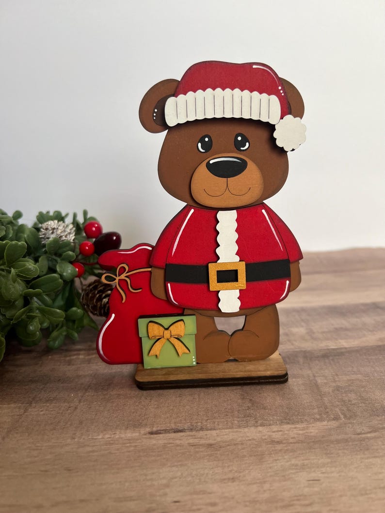 Puede incluir: Figura navide&ntilde;a de oso de madera con traje y gorro de Pap&aacute; Noel rojos con ribete blanco. El oso sostiene un saco rojo y una caja de regalo verde con un lazo dorado. La figura est&aacute; sobre una base de madera.