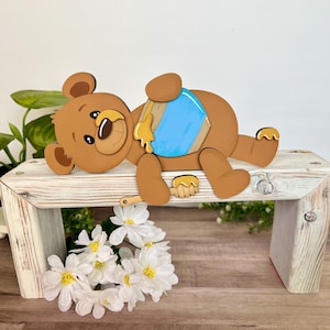 May include: A brown wooden bear with a blue diaper and honey dripping from a wooden spoon. The bear is lying on a white wooden bench with white flowers in front of it. The bear is a decorative item.