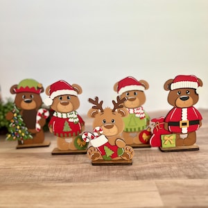Puede incluir: Una colecci&oacute;n de cinco figuras de osos navide&ntilde;os de madera. Cada oso viste atuendos festivos, incluyendo gorros de Pap&aacute; Noel, su&eacute;teres y gorros de elfo. Un oso est&aacute; dise&ntilde;ado como un reno, sosteniendo un bast&oacute;n de caramelo y un calcet&iacute;n. Las figuras est&aacute;n sobre bases de madera.