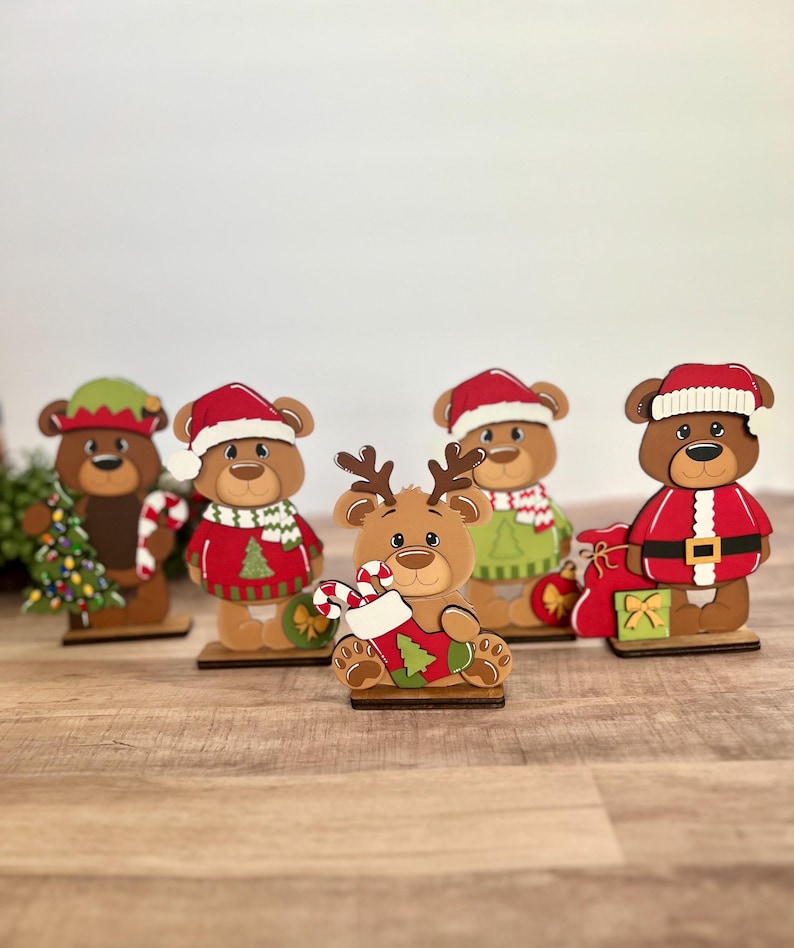 Puede incluir: Una colecci&oacute;n de cinco figuras de osos navide&ntilde;os de madera. Cada oso viste atuendos festivos, incluyendo gorros de Pap&aacute; Noel, su&eacute;teres y gorros de elfo. Un oso est&aacute; dise&ntilde;ado como un reno, sosteniendo un bast&oacute;n de caramelo y un calcet&iacute;n. Las figuras est&aacute;n sobre bases de madera.