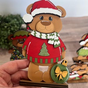 Puede incluir: Figura de oso navide&ntilde;o de madera que lleva un su&eacute;ter rojo con un dise&ntilde;o de &aacute;rbol de Navidad verde, un gorro de Pap&aacute; Noel y una bufanda verde y blanca. El oso sostiene un adorno verde con un lazo dorado. La figura est&aacute; sobre una base de madera.