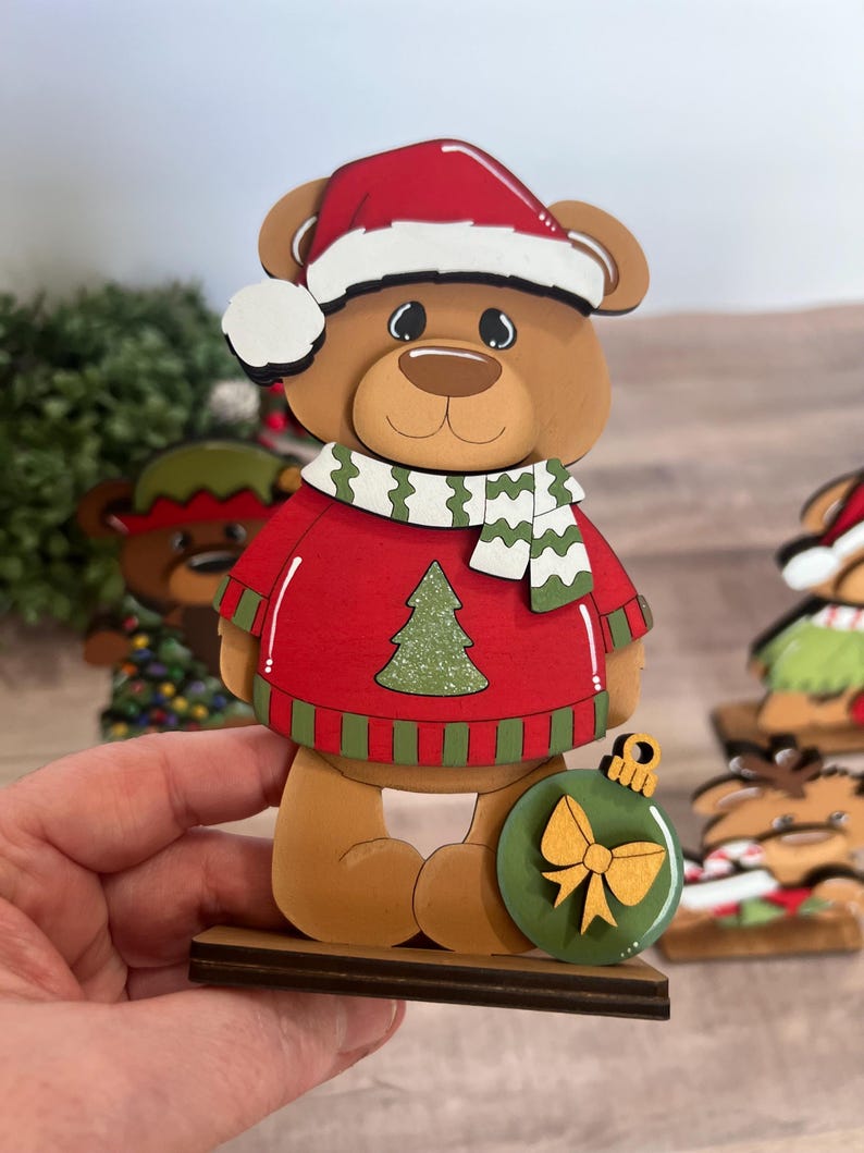 Puede incluir: Figura de oso navide&ntilde;o de madera que lleva un su&eacute;ter rojo con un dise&ntilde;o de &aacute;rbol de Navidad verde, un gorro de Pap&aacute; Noel y una bufanda verde y blanca. El oso sostiene un adorno verde con un lazo dorado. La figura est&aacute; sobre una base de madera.