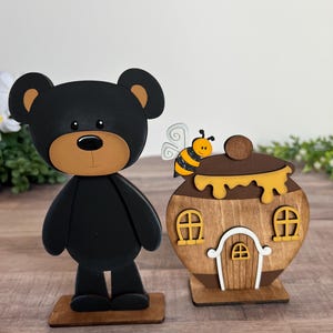 May include: A black bear figurine with a tan muzzle standing on a wooden base next to a wooden honey pot house. The honey pot features a brown finish, yellow dripping honey, two small windows, a white arched door, and a small bee perched on top.